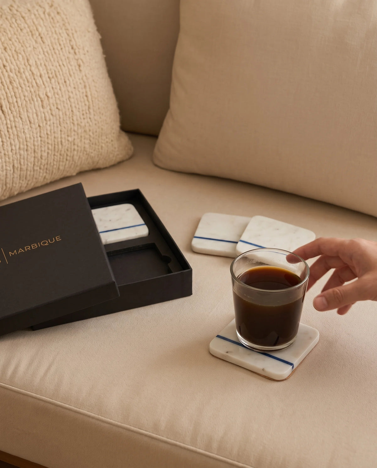 White rounded-square coasters with a slim blue stripe, styled with a coffee glass beside black packaging.