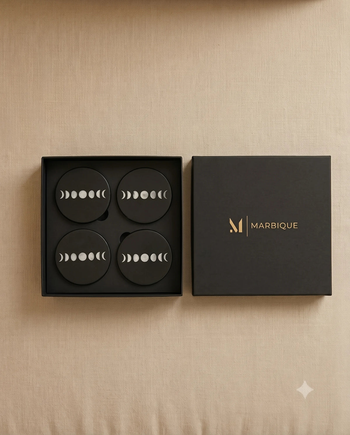 Black round coasters with pale moon-phase motifs in an open black Marbique box viewed from above.