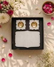 Open black box displaying a plain white shaped rectangular tray and two floral diya holders with gold-tone cups.