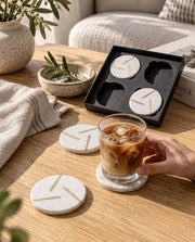 White round coasters with intersecting gold-tone lines arranged in a black Marbique box beside a matching coaster.