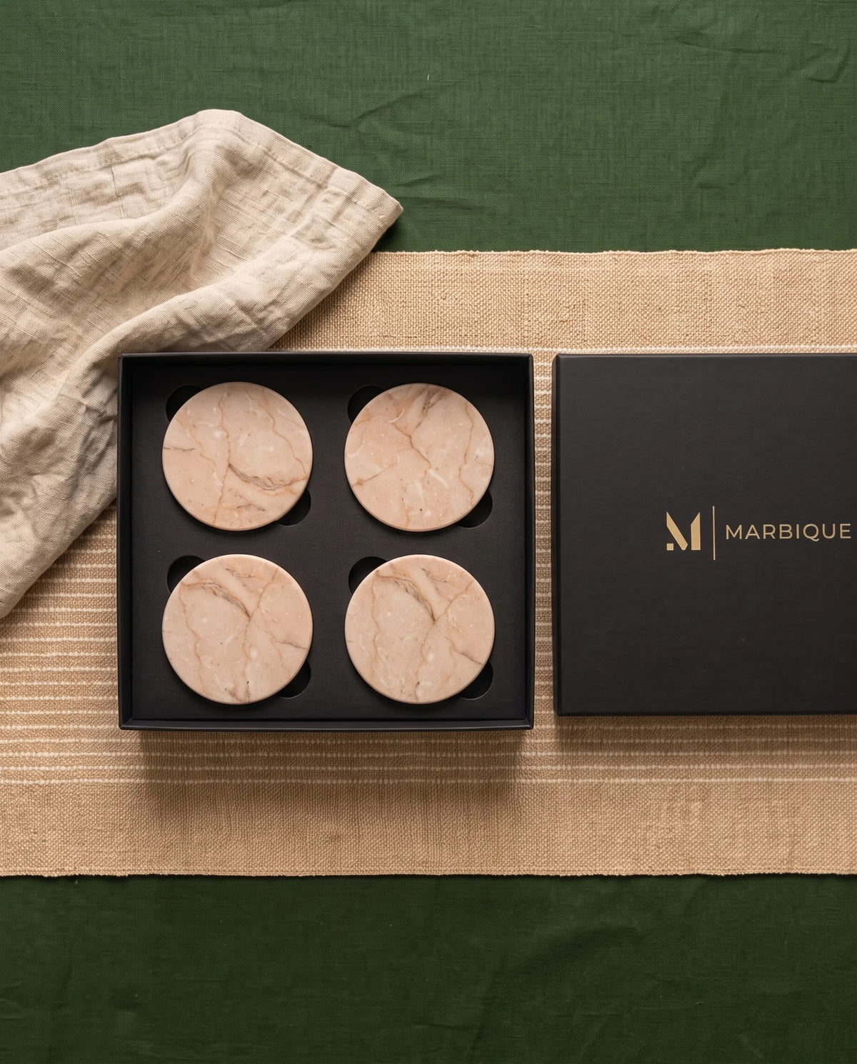 Four beige round coasters with warm tan veining arranged in an open black Marbique box.