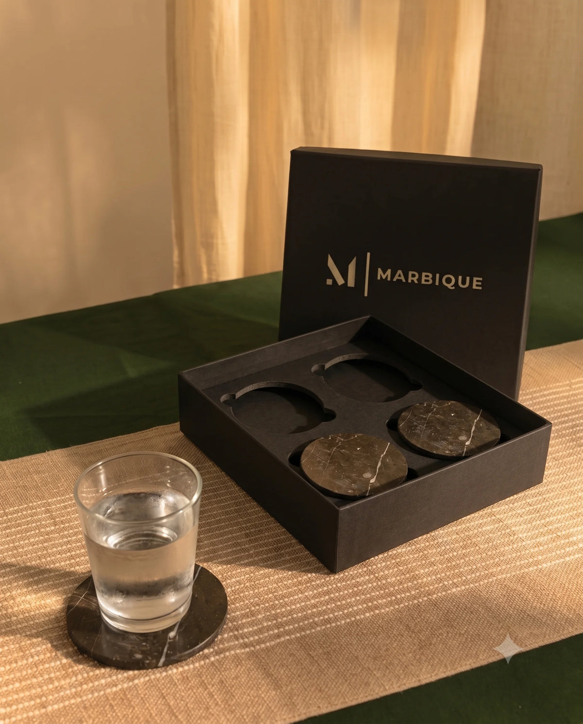 White round coasters with diagonal blue and gold-tone lines in a black Marbique box, with a drink on a matching coaster.