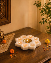 White flower-shaped holder with lit candles in petal recesses and a gold-tone central cup on a wooden altar.