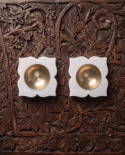 Two white four-petal diya holders with round gold-tone cups viewed from above on a carved table.