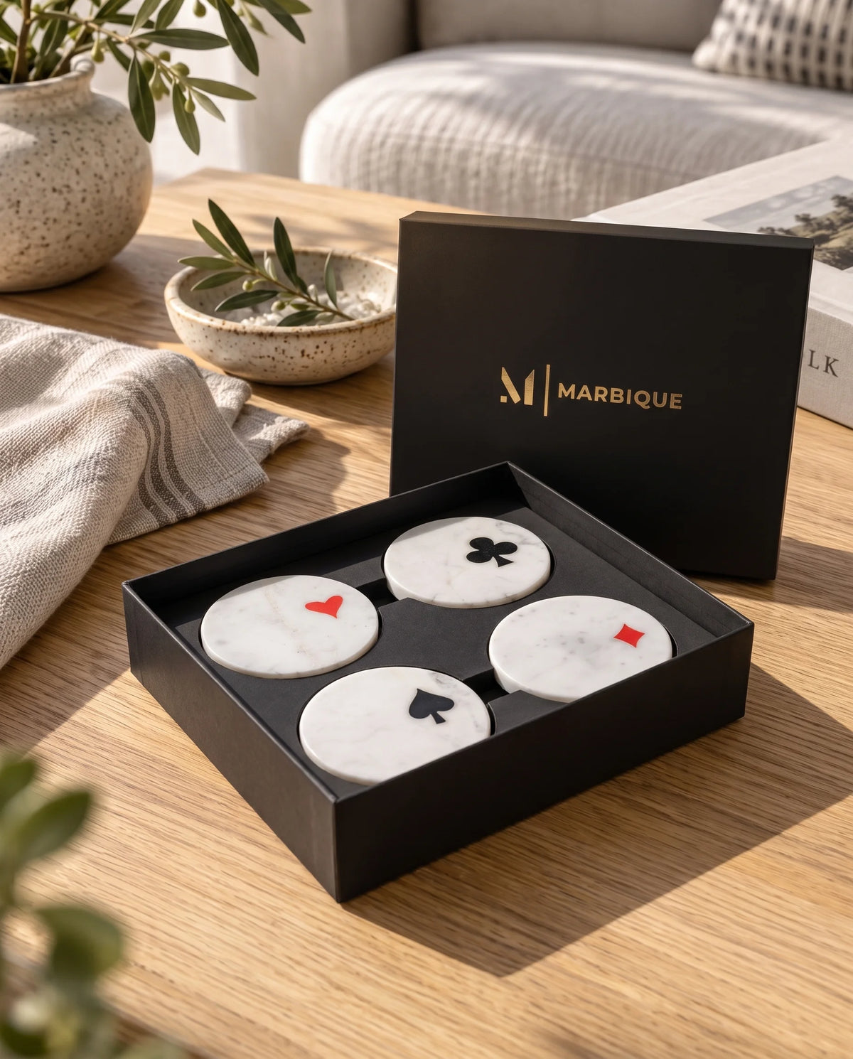 White square coasters with diagonal blue accents and gold-tone lines in a black Marbique box beside a drink.