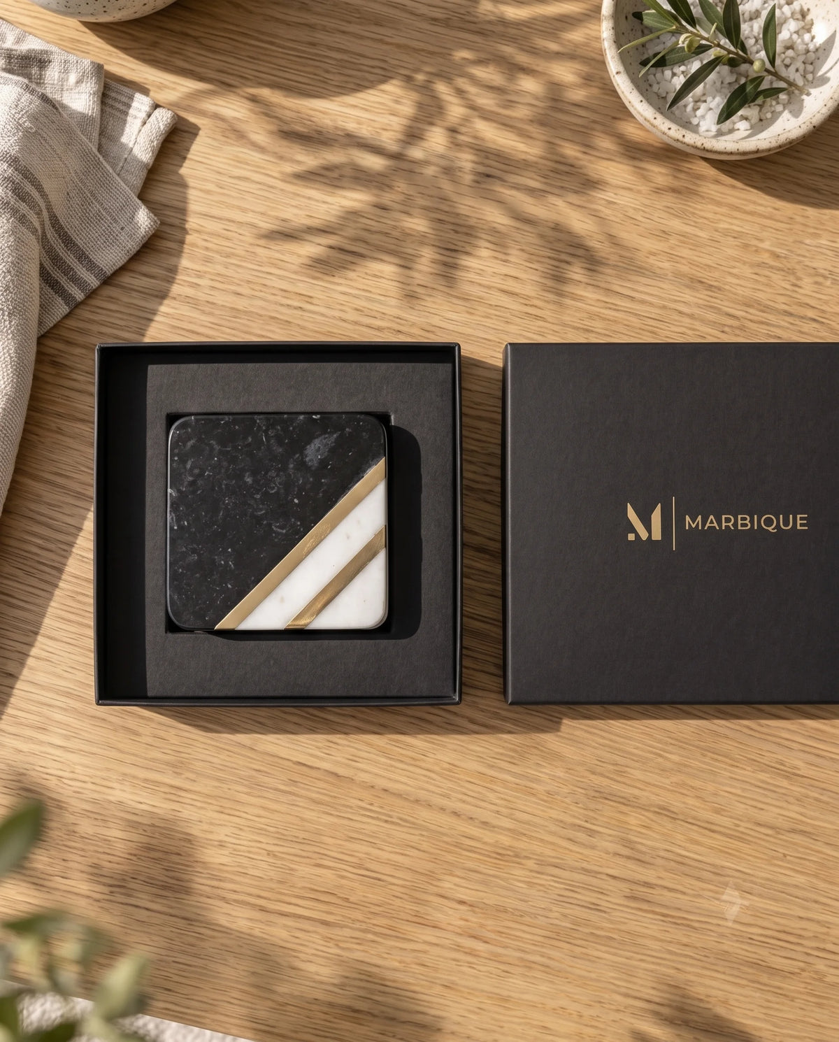 Black and white square coaster with rounded corners and diagonal gold-tone bands in an open black Marbique box.