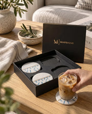 Round white coasters with blue dash details and a gold-tone divider in and beside a black Marbique box.
