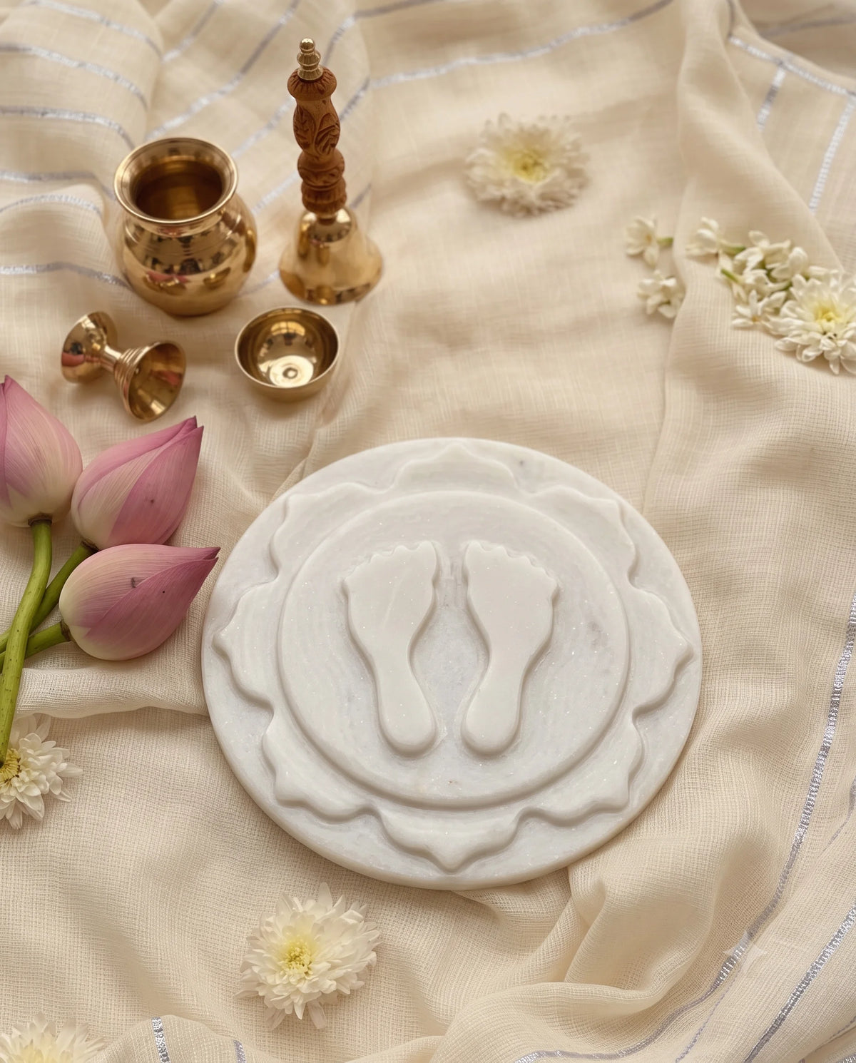 White layered footprint plaque styled with lotus buds and small gold-tone pooja vessels on cream fabric.