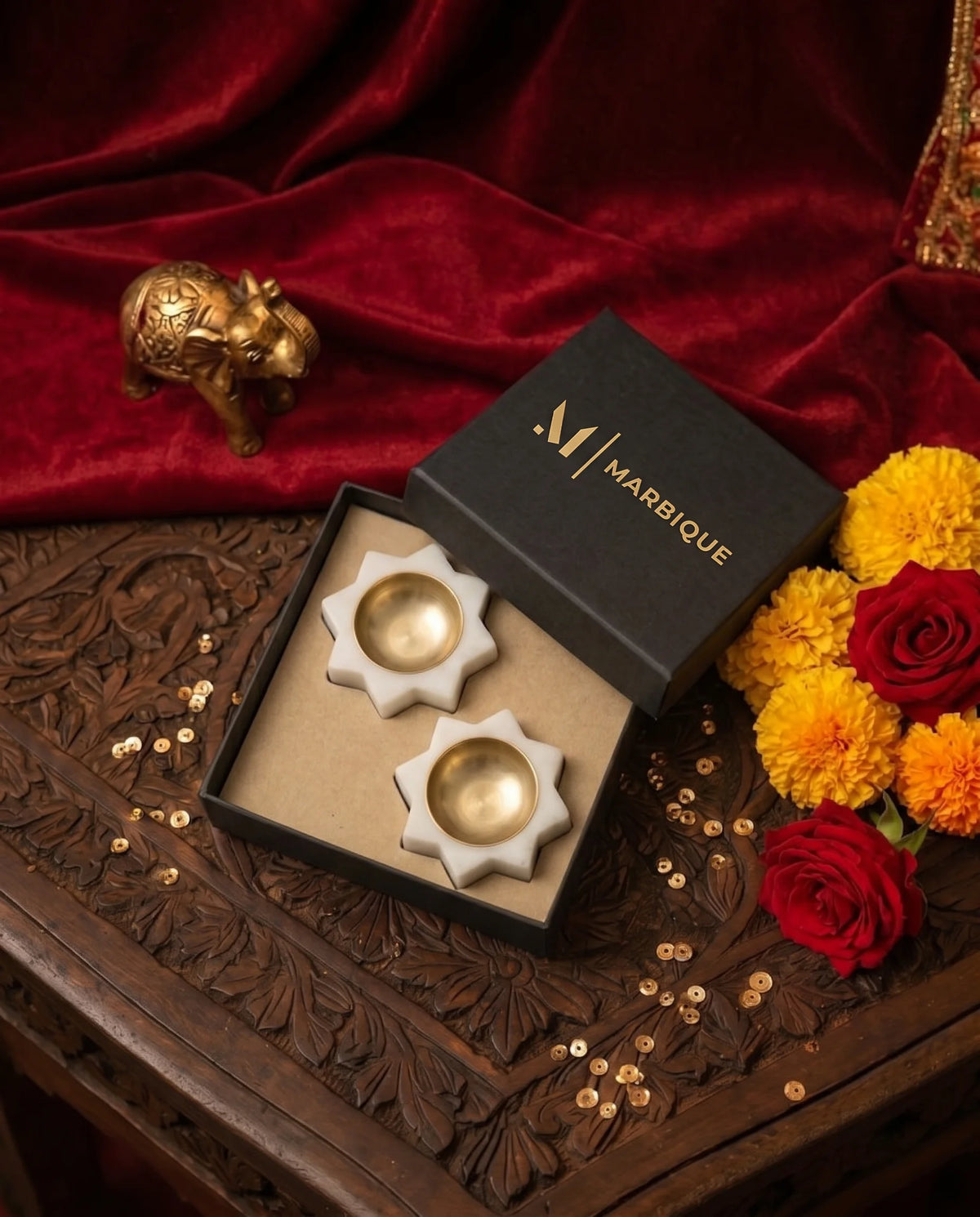 Two white pointed-star diya holders with gold-tone cups in a black Marbique box beside red fabric and flowers.