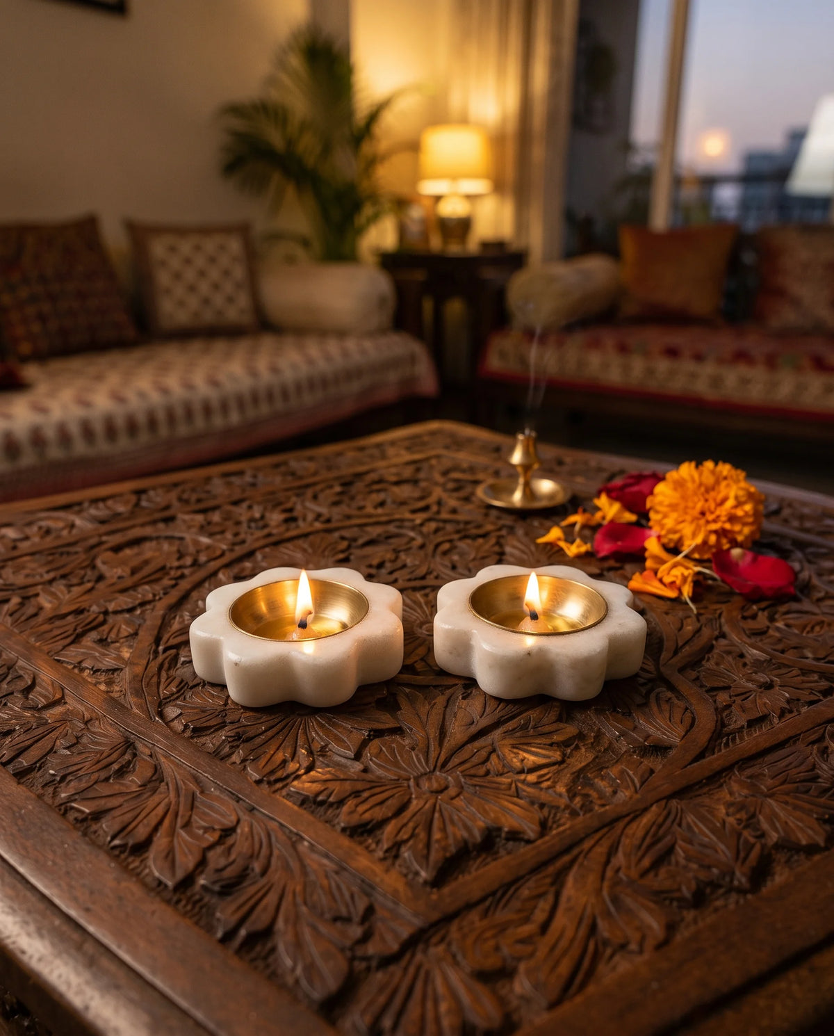 Two lit white flower-shaped diya holders on a carved table in a living-room setting.