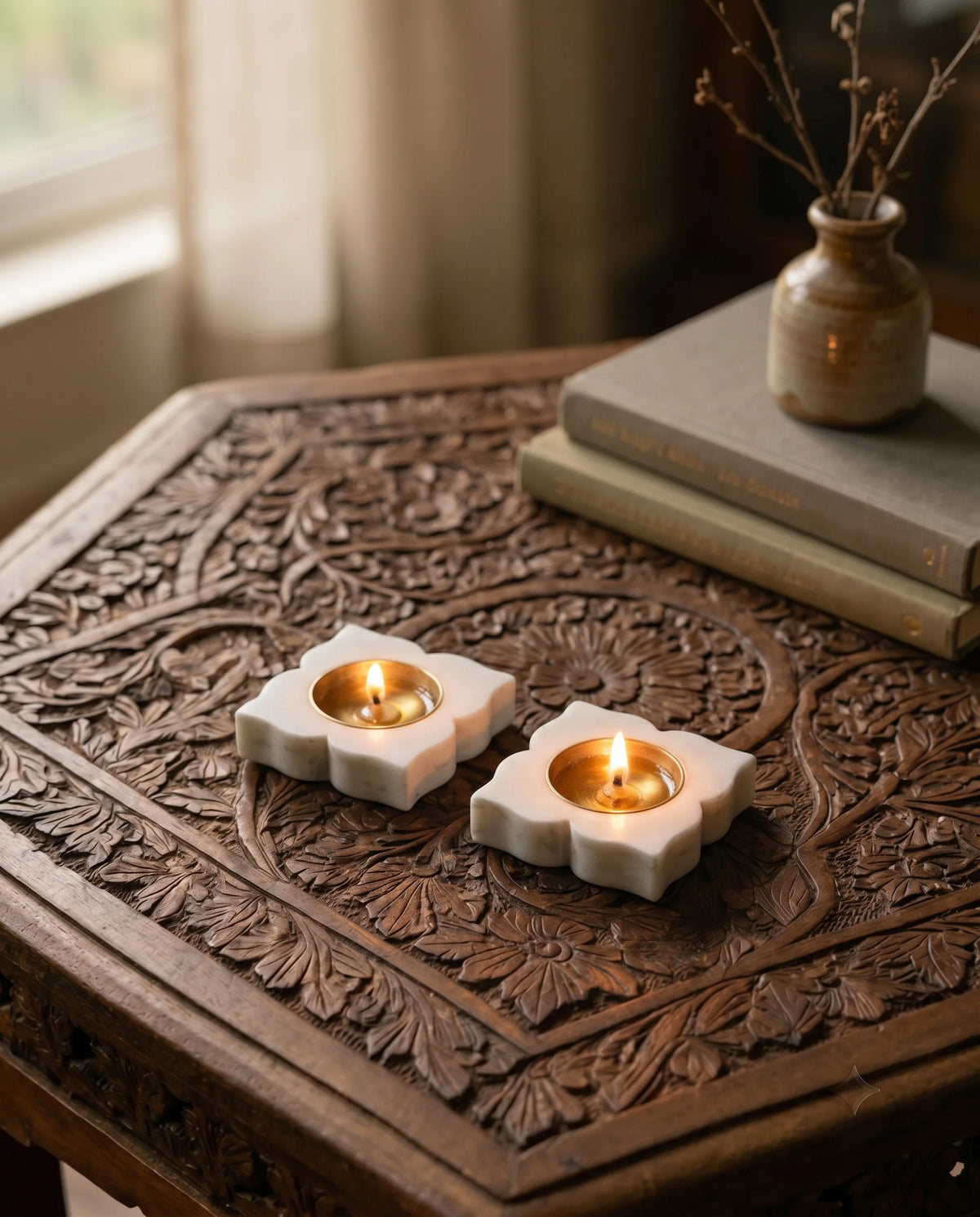 Two white four-petal diya holders with lit cups on a carved table beside books and a vase.