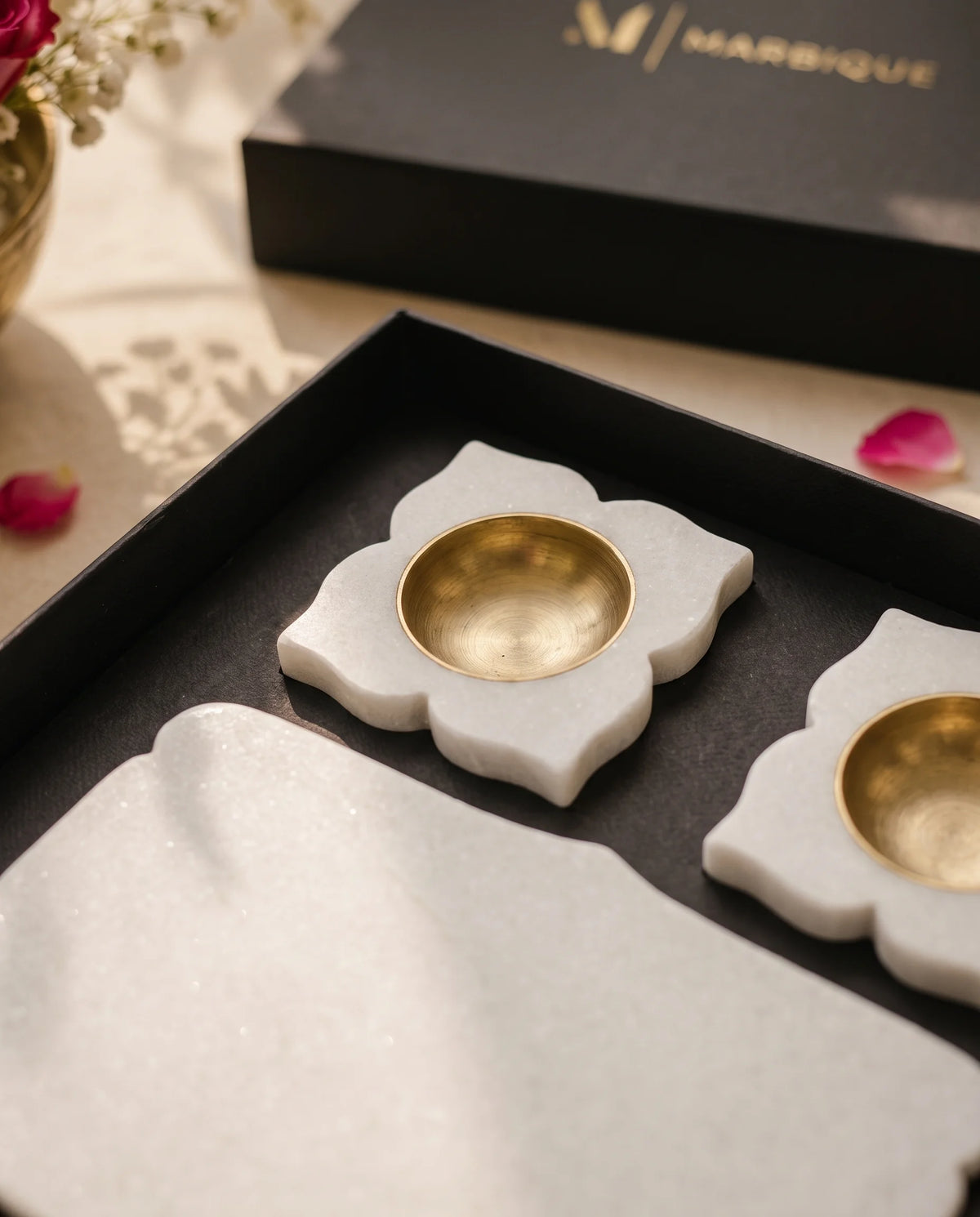 Close-up of white floral diya-holder designs with round gold-tone cups beside the shaped white tray in a black box.