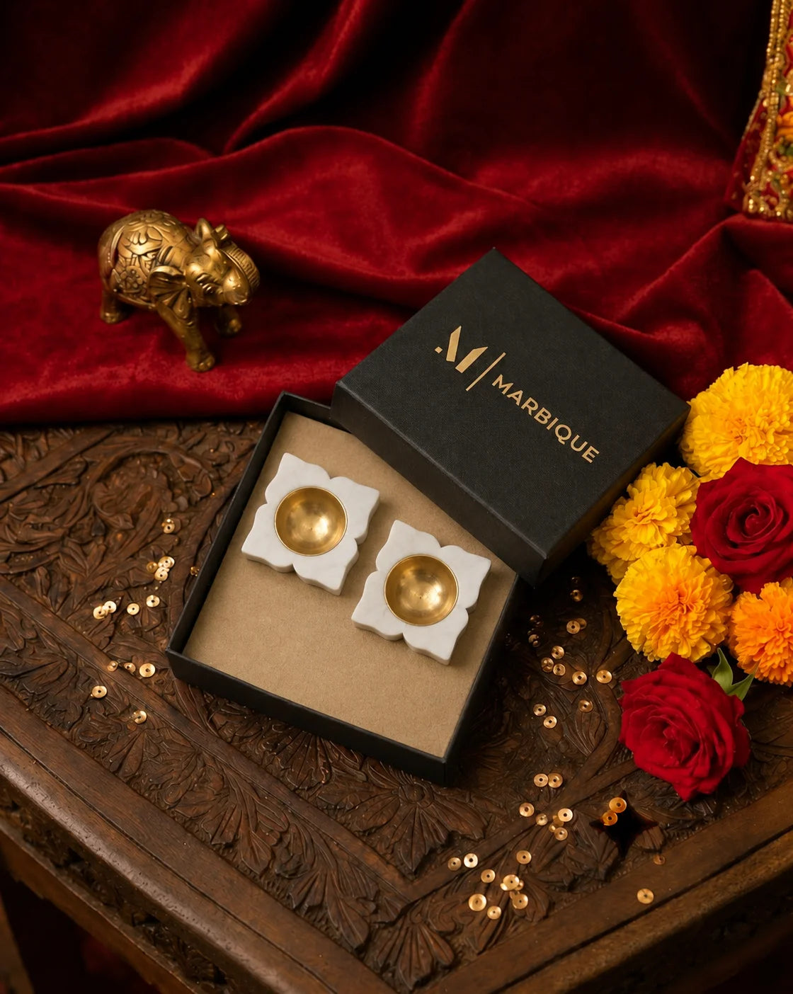 Two white floral diya holders with gold-tone cups in an open black Marbique box beside flowers and red fabric.