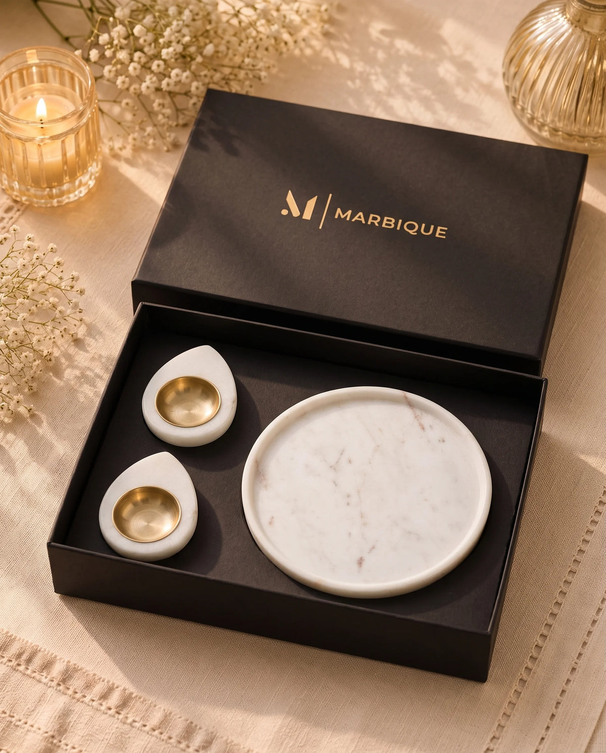 White round dish and teardrop holders styled in their box beside a decorative candle and flowers.