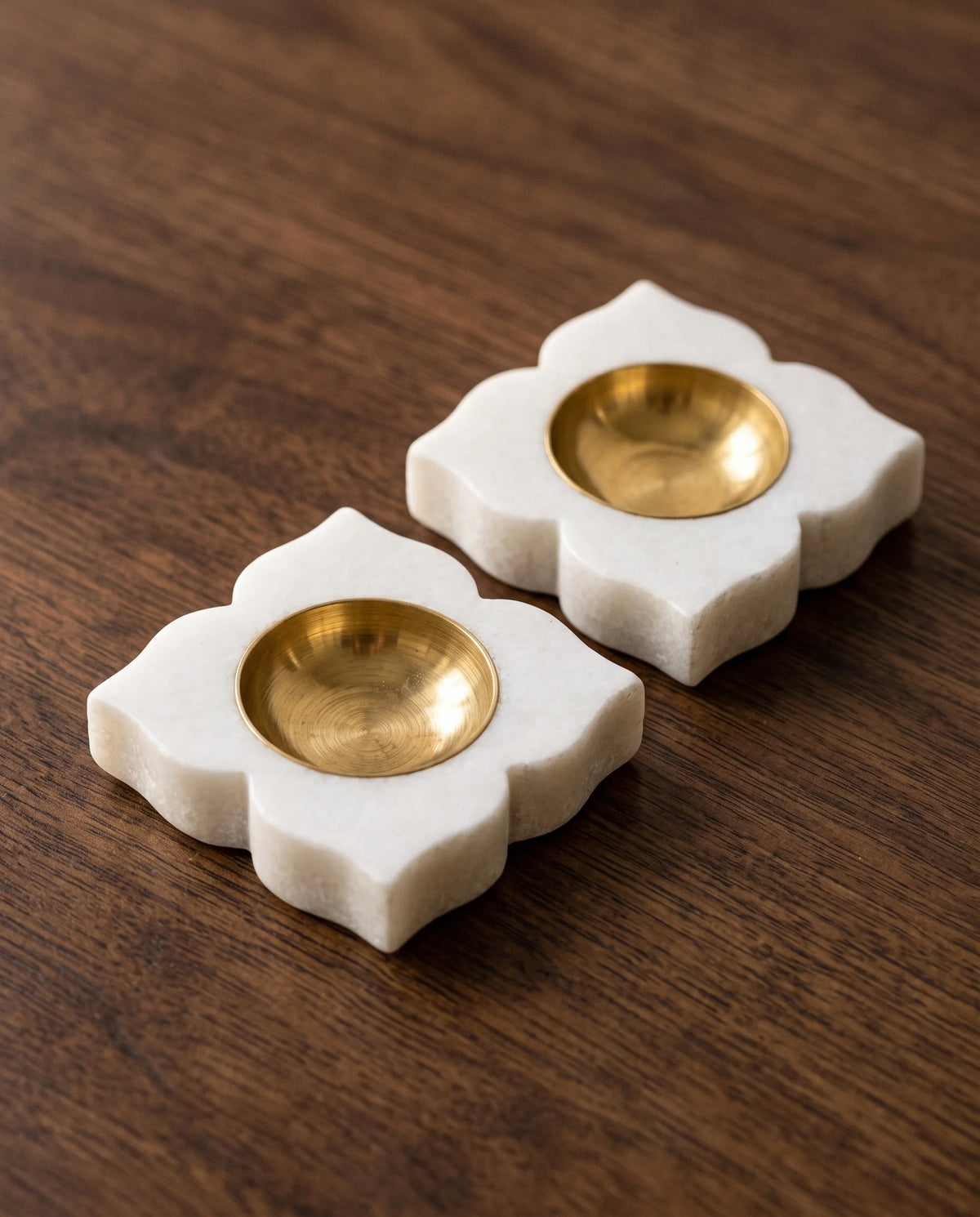 Close view of two white four-petal diya holders with round gold-tone cups on a wooden surface.