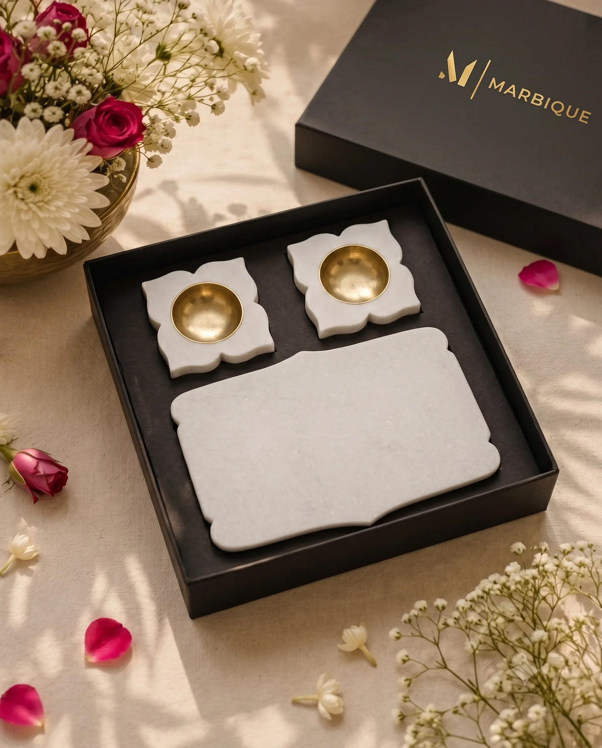 Angled view of a plain white shaped tray and two floral diya holders in a black Marbique box beside flowers.