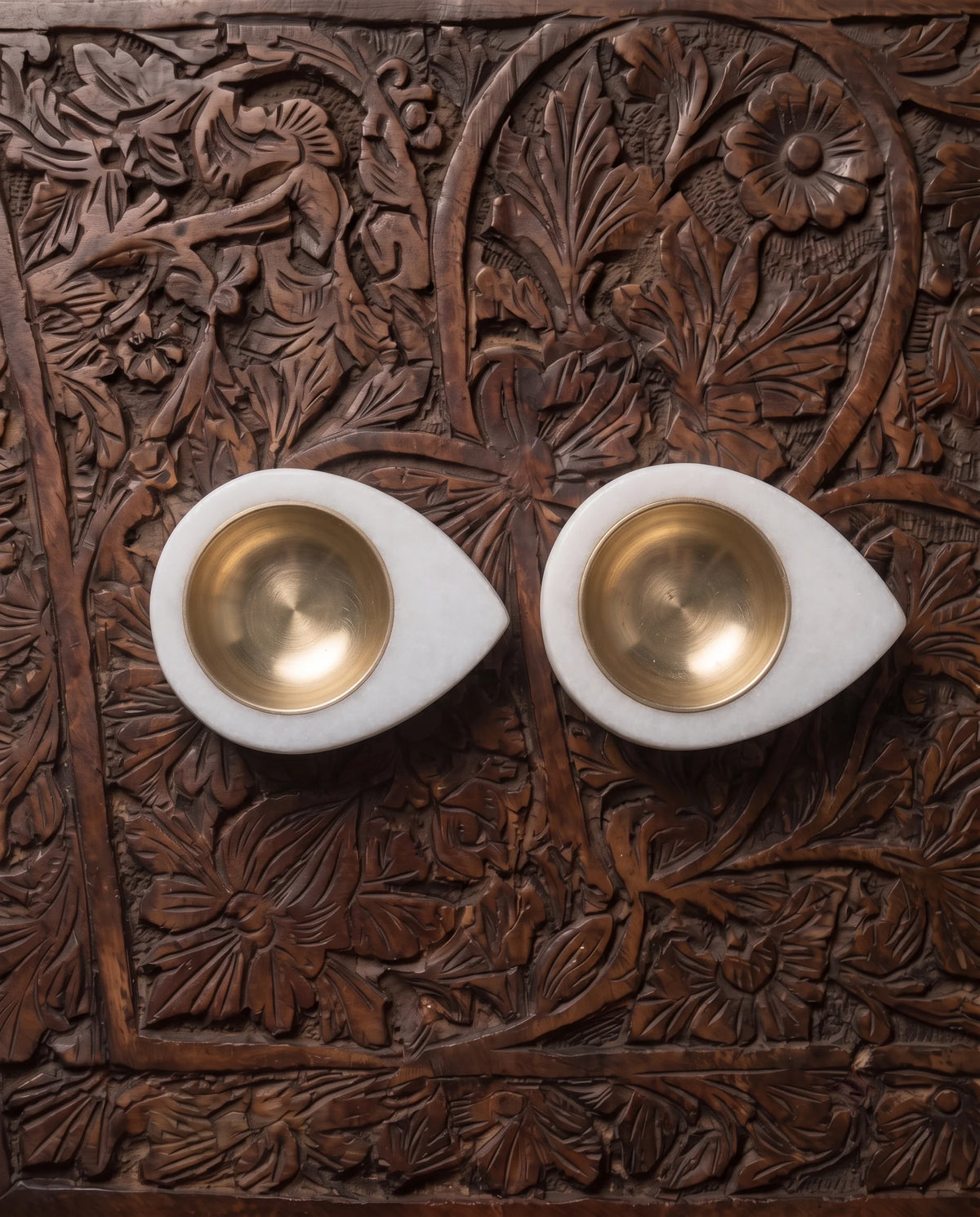 Overhead view of two matching white teardrop diya holders with gold-tone cups on a carved surface.