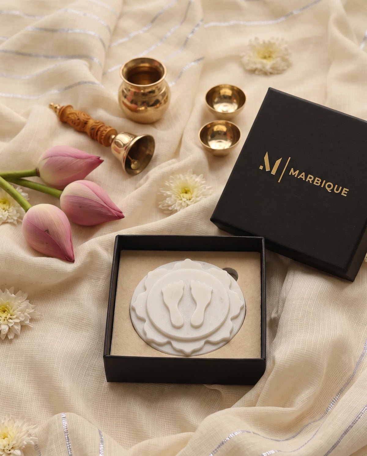 White layered footprint plaque displayed in a black Marbique box, with pooja vessels and lotus buds outside.