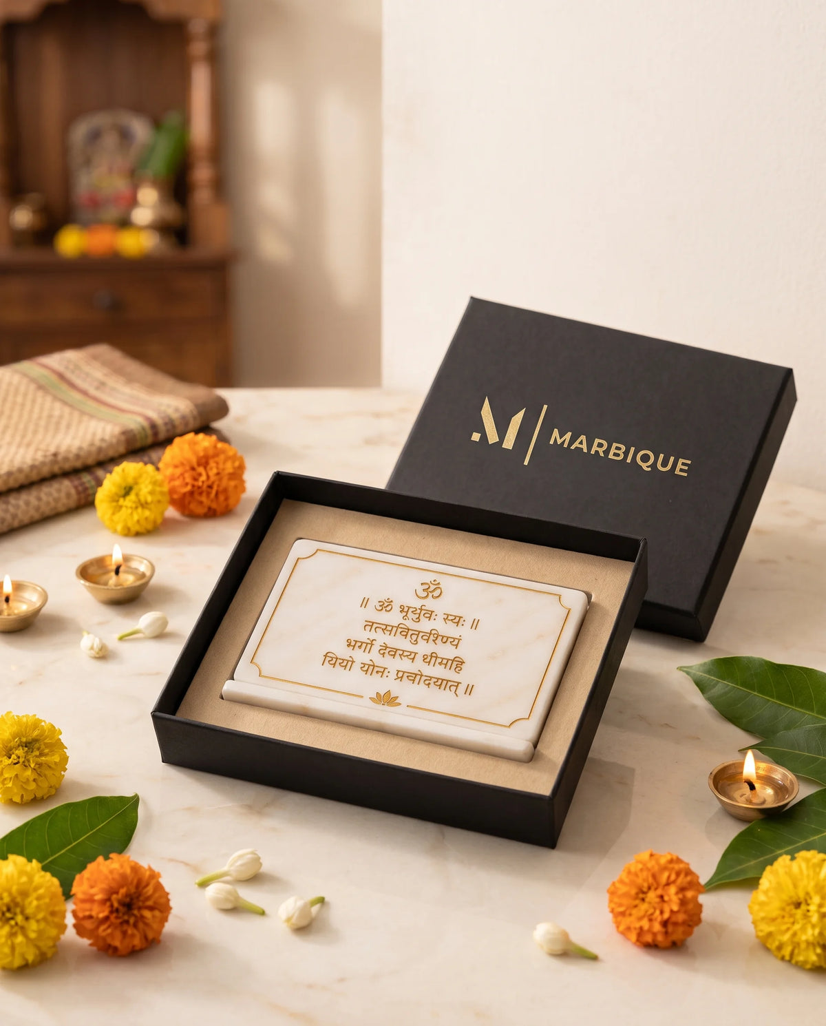 White Gayatri Mantra plaque displayed flat in an open black Marbique box, with flowers and lamps outside.