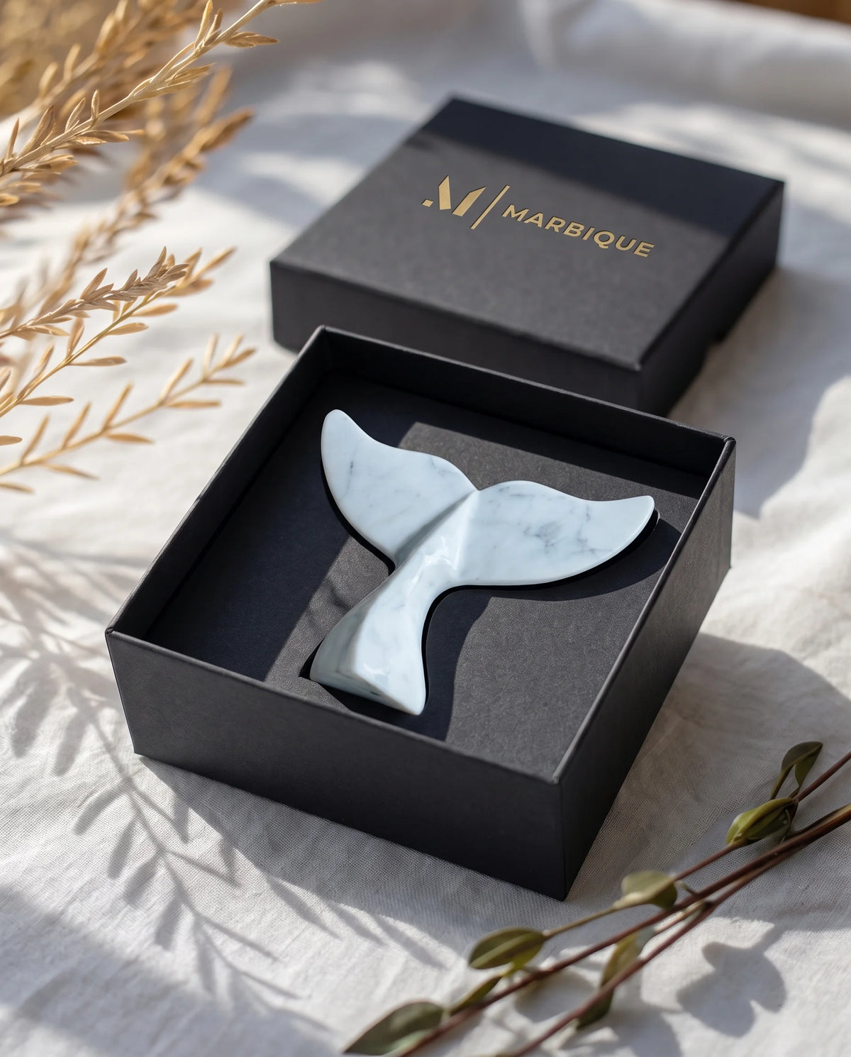 White whale-tail sculpture with grey veining in an open black Marbique box on light fabric.