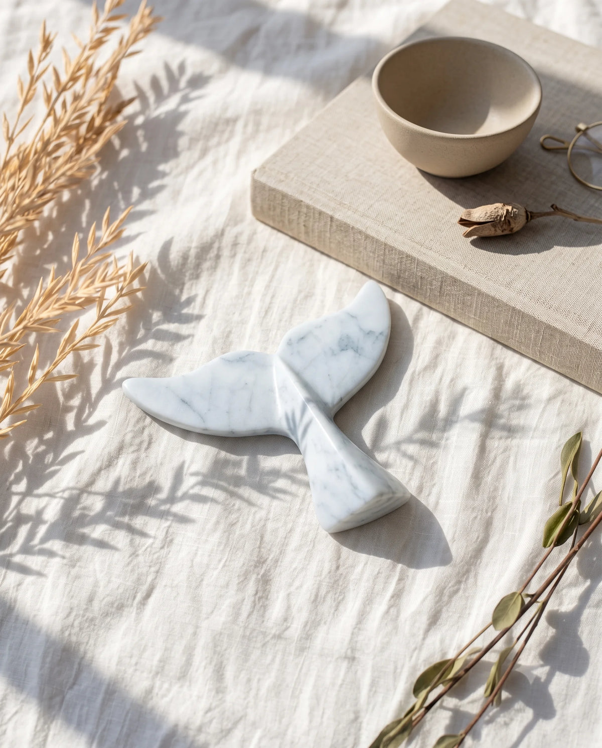 White whale-tail decorative form with grey veins styled on fabric beside a book, bowl and glasses.