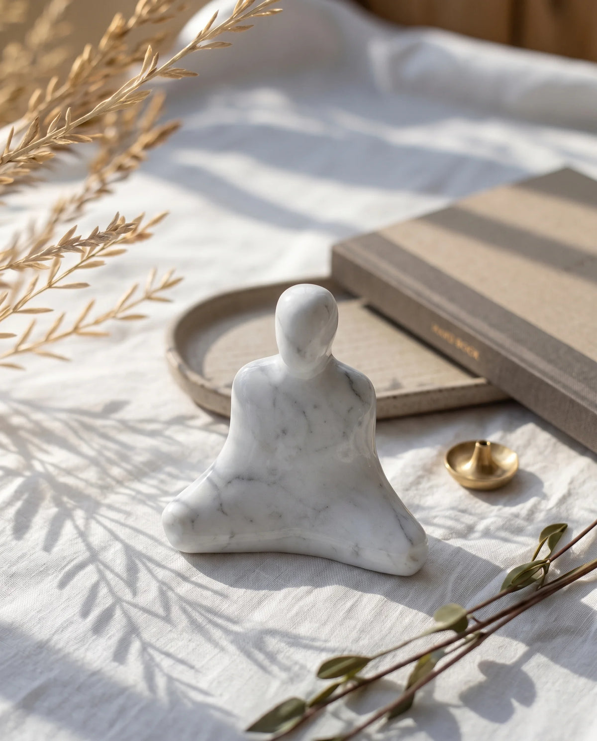 White seated-figure sculpture with grey veining on light fabric beside books, a dish and a small gold-tone object.