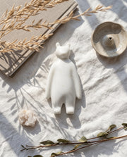 White bear-like decorative figure with rounded ears and long arms on fabric beside books, dried stems and a bowl.