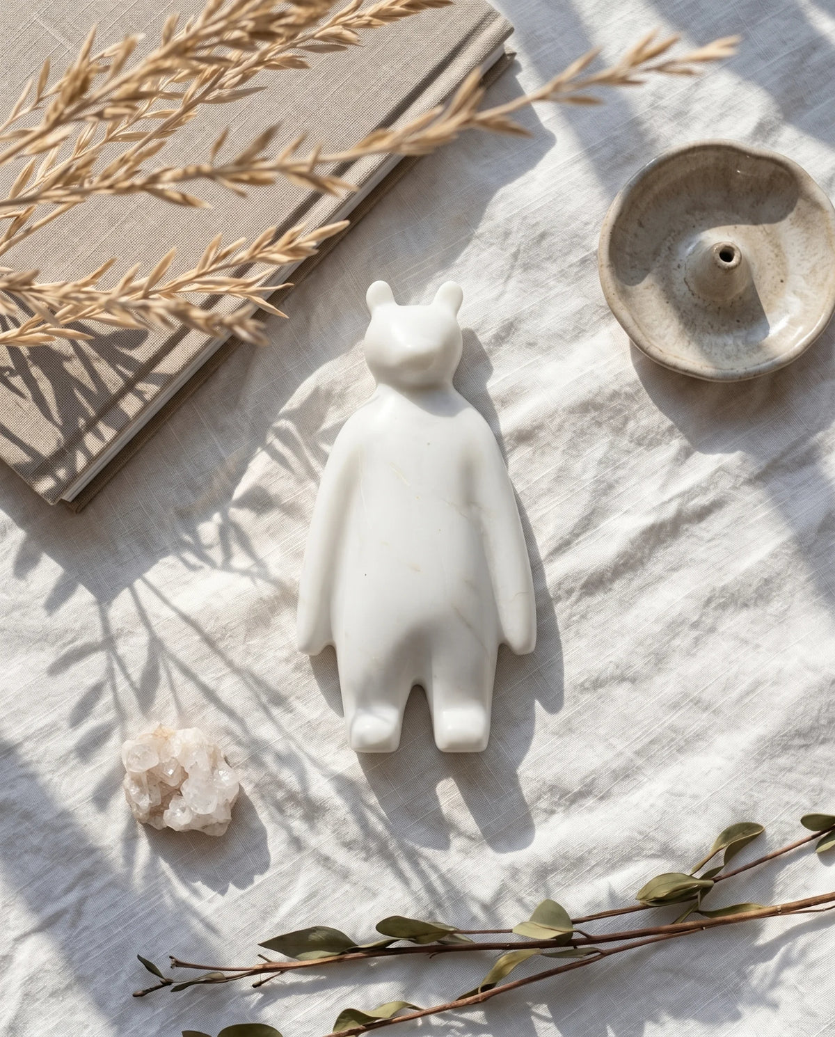 White bear-like decorative figure with rounded ears and long arms on fabric beside books, dried stems and a bowl.