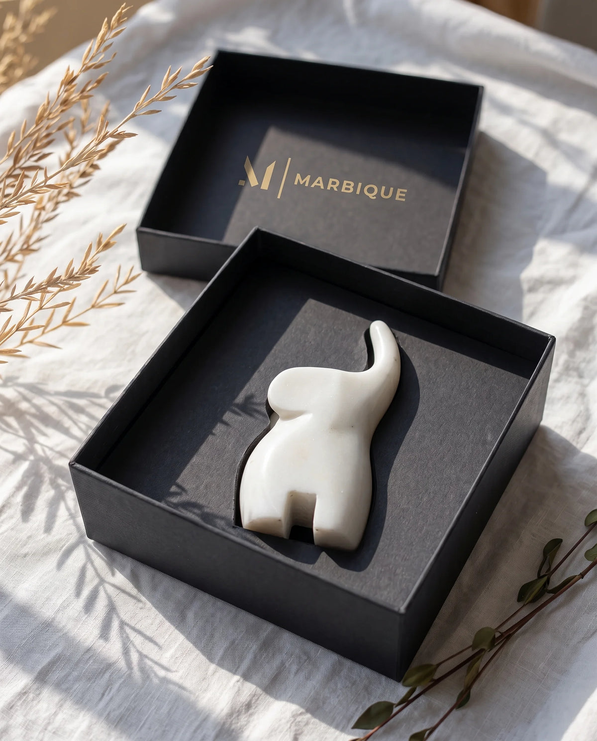 White raised-trunk elephant sculpture in an open black Marbique box with the lid behind.