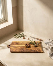 Brown shaped rectangular board with a short handle, styled with olives and leaves beside a knife and garlic.