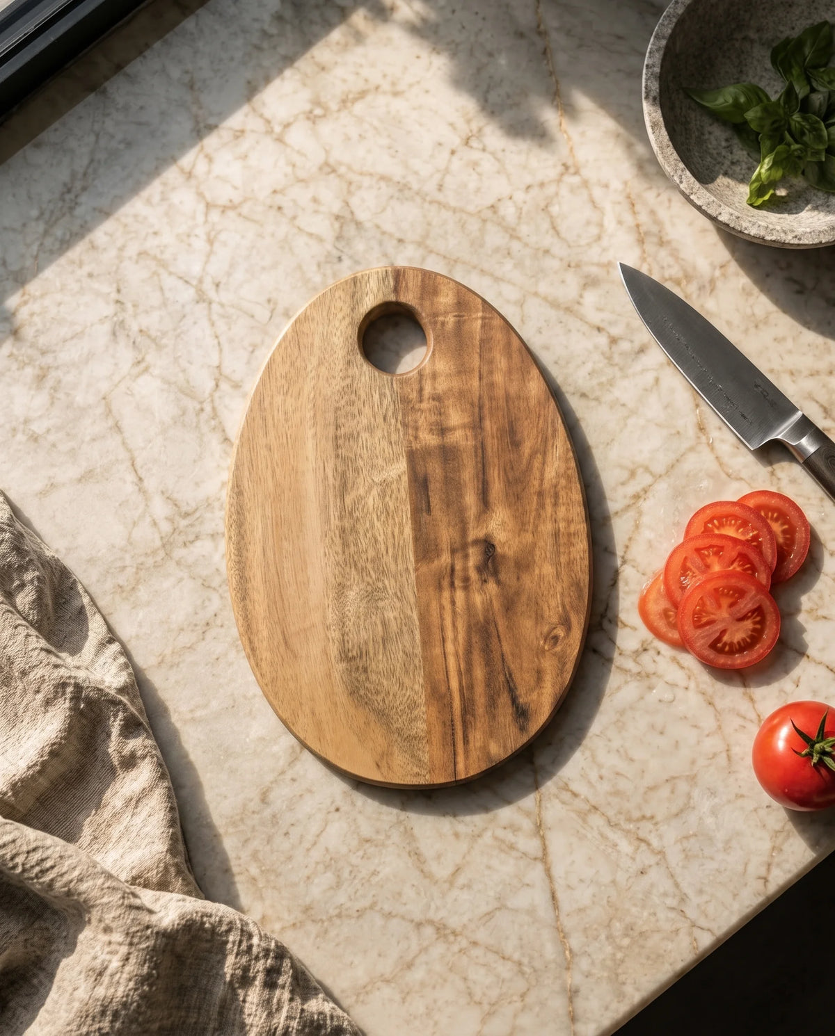 Overhead view of a plain two-tone oval board beside sliced tomatoes, a knife and fabric.
