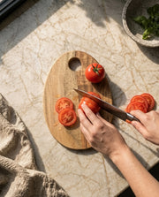 Hands shown slicing a tomato on the two-tone oval board beside a circular opening.