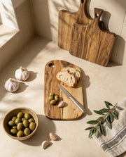 Brown rounded rectangular board with bread, olives and a knife on a counter, with two other boards leaning behind.