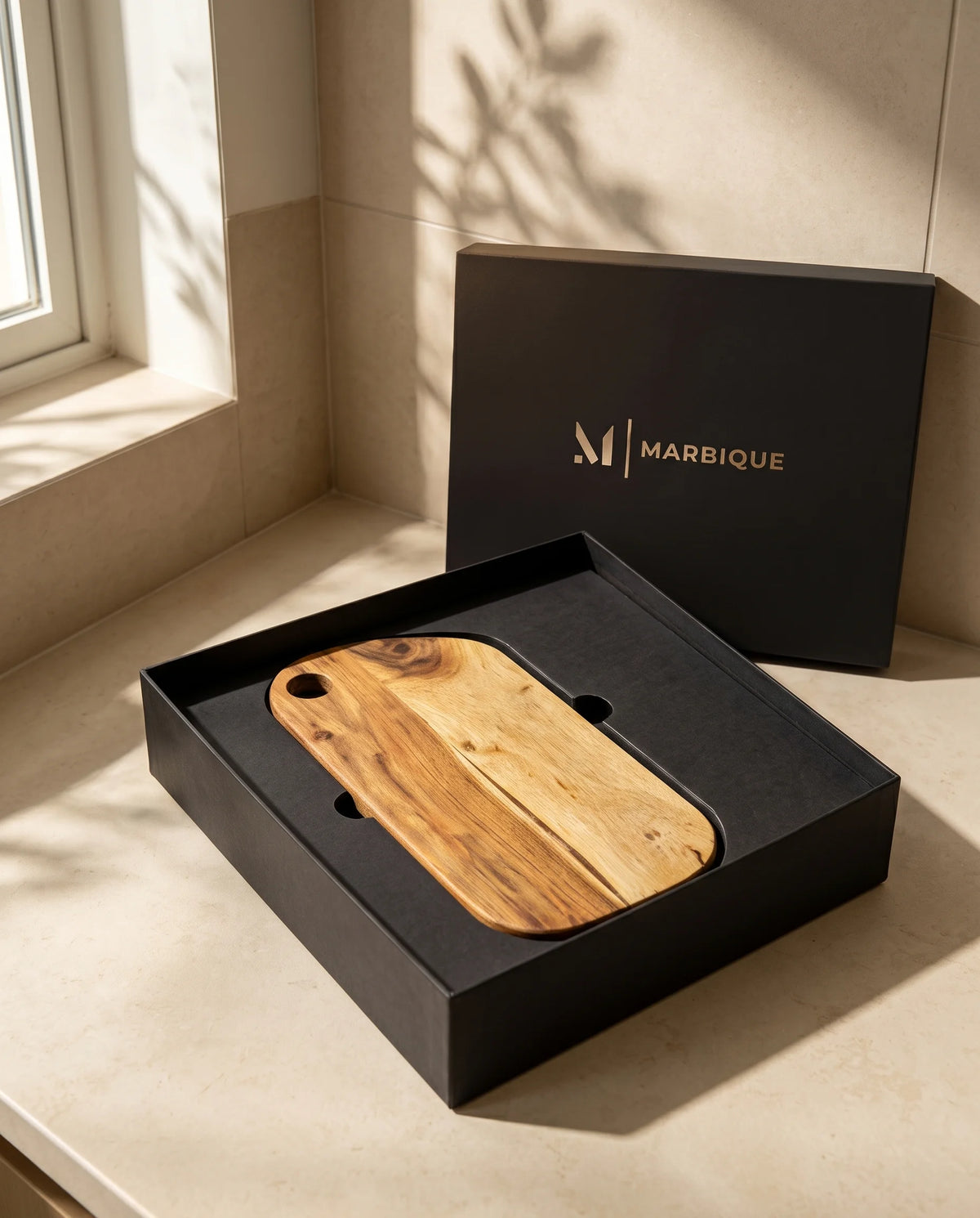Brown rounded rectangular corner-hole board displayed in an open black Marbique box beside a window.
